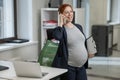 Pregnant woman using mobile phone and holding paper tablet in office. Royalty Free Stock Photo