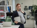 Pregnant woman using mobile phone and holding paper tablet in office. Royalty Free Stock Photo