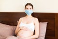 Pregnant woman is wearing protective medical mask with word. Mother is staying in bed because of respiratory disease.