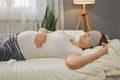 Pregnant woman wearing white casual T-shirt and blindfold sleeping in bed at home in bedroom interior touching her tummy resting