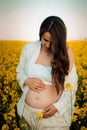 Pregnant woman in white clothes hugs her belly in a rapeseed field