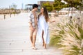 Pregnant woman in white dress embraced with handsome man wearing unbuttoned shirt walking on the beach sidewalk path. Royalty Free Stock Photo