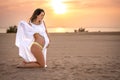 Pregnant woman in white dress standing on knees, holding her stomach and enjoying ocean beach coast at sunset. Royalty Free Stock Photo