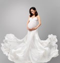 Pregnant Woman in White Long Dress flying on Wind looking at Belly. Beautiful Mother in Silk waving Gown over Gray Background. Royalty Free Stock Photo