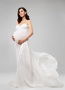 Pregnant Woman in White Long Dress flying on Wind looking up. Beautiful Mother dreaming in Silk waving Gown over Gray Background.