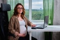 Pregnant woman working at a laptop in a home office in the background of a window Royalty Free Stock Photo