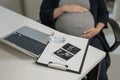 A pregnant woman works on a laptop in the office and looks at a photo from an ultrasound scan of the fetus. Belly close Royalty Free Stock Photo