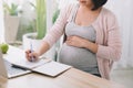 Closeup of a pregnant woman writing notes and using a laptop while working on maternity leave at her dining room table at home Royalty Free Stock Photo