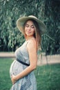 Pregnant young Caucasian woman wearing long blue dress and rustic country hat in park outside. Royalty Free Stock Photo