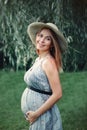 Pregnant young Caucasian woman wearing long blue dress and rustic country hat in park outside. Royalty Free Stock Photo