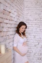 Pregnant young woman wearing white dress standing in brick wall background and holding belly.