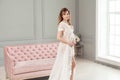 Pregnant young woman in white dress peignoir standing near pink sofa, holding with love her belly Royalty Free Stock Photo