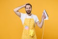 Preoccupied puzzled young man househusband in apron hold in hands iron while doing housework isolated on yellow