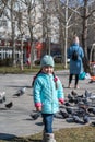 A preschool-aged girl in a blue jacket poses outside in front of a flock of city pigeons. A happy five-year-old child with long