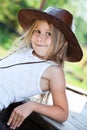 Preschool blond hair girl dressed sleeveless sports shirt and brown cowboy hat sitting on wooden veranda of timber house