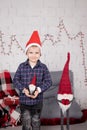 preschool boy in santa hat standing near tall christmas gnome toy, holding little dwarf toy in hand. in grey room. happy Royalty Free Stock Photo
