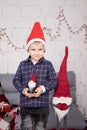 preschool boy in santa hat standing near tall christmas gnome toy, holding little dwarf toy in hand. in grey room. happy Royalty Free Stock Photo