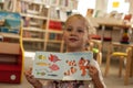 Preschooler little girl sitting and reading a book in library. Kid with books near a bookcase. Happy, cheerful and cute girl read