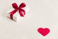 Present for Valentine`s day, Mother`s day or Wedding, wrapped in white paper, red bow next to a red heart on a light textile backg