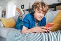 Preteen boy lying at home on cozy sofa dressed casual jeans and shirt. listening to music and chatting using wireless headphones Royalty Free Stock Photo