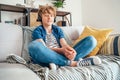 Preteen boy sitting cross-legged at home on cozy sofa dressed casual jeans and new sneakers listening to music using wireless