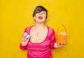 Pretty adult chubby woman with short hair holding colorful holiday eggs and a easter basket on yellow solid studio background