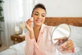 Pretty african american lady applying cosmetic foundation with applicator, holding mirror, sitting on bed in bedroom Royalty Free Stock Photo