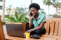 A pretty African woman is sitting at a table in a street cafe, talking on the phone and using a laptop. A young woman is Royalty Free Stock Photo