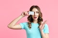 Pretty amazed female holding credit card at her face closing eyes isolated over pink background