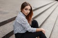 Pretty American young woman in fashionable jeans in a vintage black and white striped blouse with elegant velvet necklaces sits