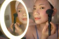 Pretty Asian woman brushing her face - lifestyle portrait of young happy and beautiful Japanese girl at home applying makeup and Royalty Free Stock Photo