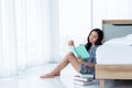 Pretty Asian woman sitting near a white bed in her bedroom, She is resting by reading a notebook Which recorded her