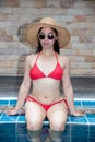 Pretty Asian woman wearing red bikini sitting at swimming pool edge with straw hat and sunglasses. Royalty Free Stock Photo