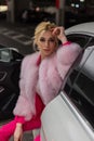 Pretty attractive young woman with blond hair in fashionable glamorous pink fur coat is sitting in luxurious white car. Beautiful Royalty Free Stock Photo