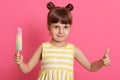 Pretty baby girl kid eating ice cream, looks happy and showing thumb up, posing isolated over pink background, wearing white and Royalty Free Stock Photo