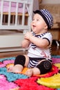 A pretty baby in a striped shirt and hats seated on the mat in the room
