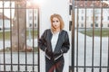 Pretty beautiful young blond woman in a black spring stylish jacket in black jeans in white shirt stands near the gate