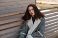 Pretty beautiful young brunette woman in a stylish plaid jacket with white fur is sitting and relaxes on a bench in the city