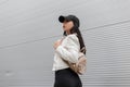 Pretty beautiful young woman in a fashionable white leather jacket in stylish black jeans with a trendy gold backpack walks Royalty Free Stock Photo