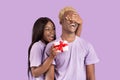 Pretty black woman covering her boyfriend`s eyes, surprising him with Valentine`s gift on lilac studio background