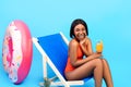 Pretty black woman drinking tropical cocktail in lounge chair, laughing, wearing stylish bikini over blue background Royalty Free Stock Photo