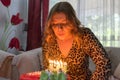 Pretty blond fashion girl blowing the candles of her birthday cake, sitting in a sofa. Happy teenager with curly hair and makeup