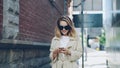 Pretty blond girl is using modern smartphone touching screen walking in city and smiling. Young woman is wearing Royalty Free Stock Photo