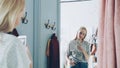 Pretty blond woman is making mirror selfie with smart phone while standing in fitting room in clothes boutique. She is Royalty Free Stock Photo