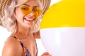 Pretty blonde caucasian female stands in swimsuit with big rubber beach colourful ball and smiles isolated on white