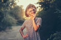 Pretty blonde girl with short haircut is posing and smiling to the camera in forest on sunset background. She wears hat Royalty Free Stock Photo