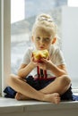 Pretty blonde little girl sitting on the sill at home, looking in window and holding red apple. Healthy food concept Royalty Free Stock Photo