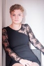 Pretty blonde ukrainian young woman with short hair sitting on a chair, dressed with black dress, against light background Royalty Free Stock Photo