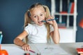 Pretty blondy girl thinking while doing her homework and holding a blue pencil, at home table. Education and school concept