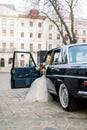 Pretty bride with wedding bouquet in luxury wedding dress sitting in the retro car, parked in old city center Royalty Free Stock Photo